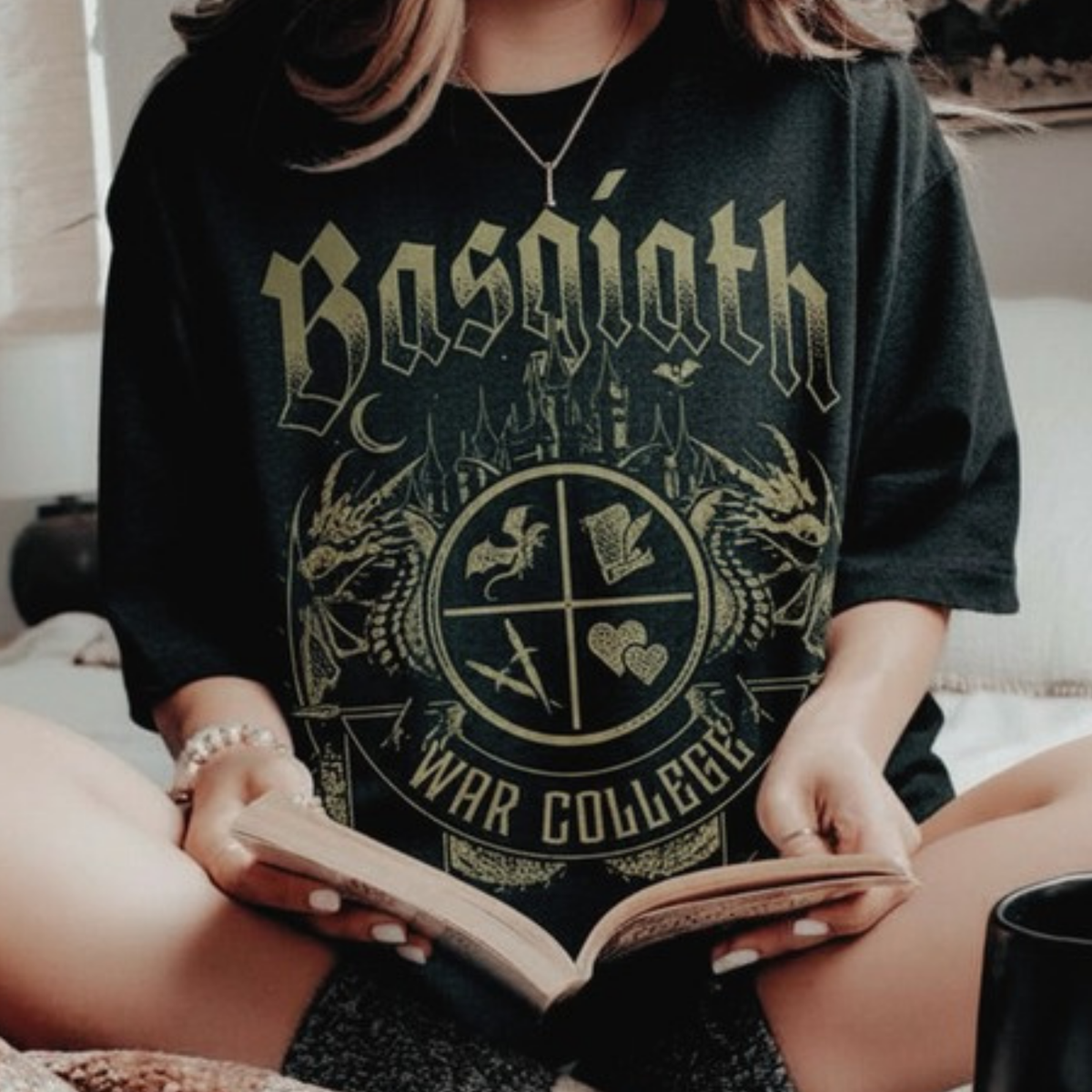 Reader holding an open book in the black Basgiath War College t-shirt, gold War College crest with dragons and castle, Fourth Wing merch inspired by Rebecca Yarros