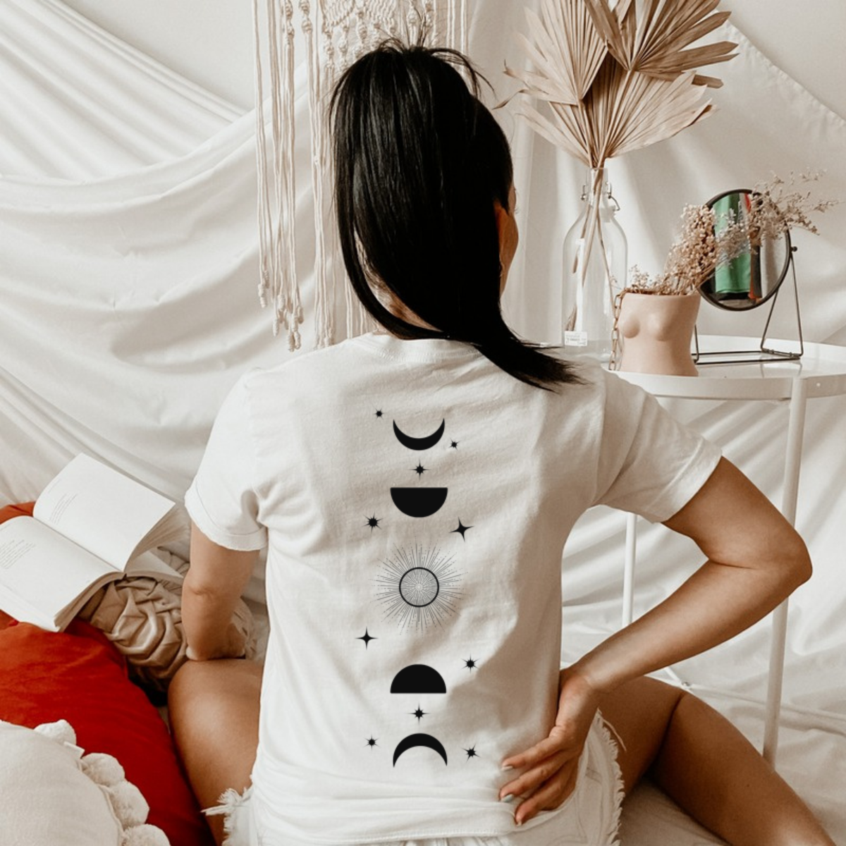 Model seated on a bed in a white Feyres Tattoo t-shirt showing the moon phase back print, ACOTAR merch inspired by Sarah J. Maas
