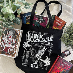 Flat-lay of a black Manon Blackbeak tote bag with a white wyvern gig-poster print, styled with Sarah J. Maas hardbacks, inspired by Throne of Glass