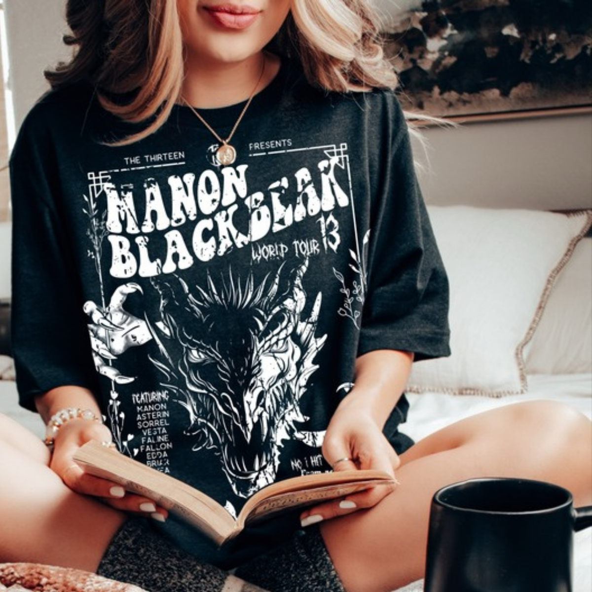 Model reading in bed wearing the black Manon Blackbeak World Tour t-shirt, inspired by Throne of Glass by Sarah J. Maas