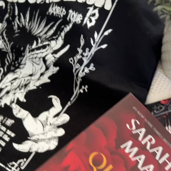 Product video: the black Manon Blackbeak tote with the Sarah J Maas hardbacks — the white line-art wyvern skull under 'MANON BLACKBEAK / WORLD TOUR 13' in the concert-poster layout [focal 50 55]