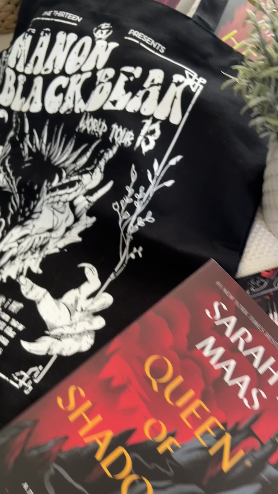 Product video: the black Manon Blackbeak tote with the Sarah J Maas hardbacks — the white line-art wyvern skull under 'MANON BLACKBEAK / WORLD TOUR 13' in the concert-poster layout [focal 50 55]