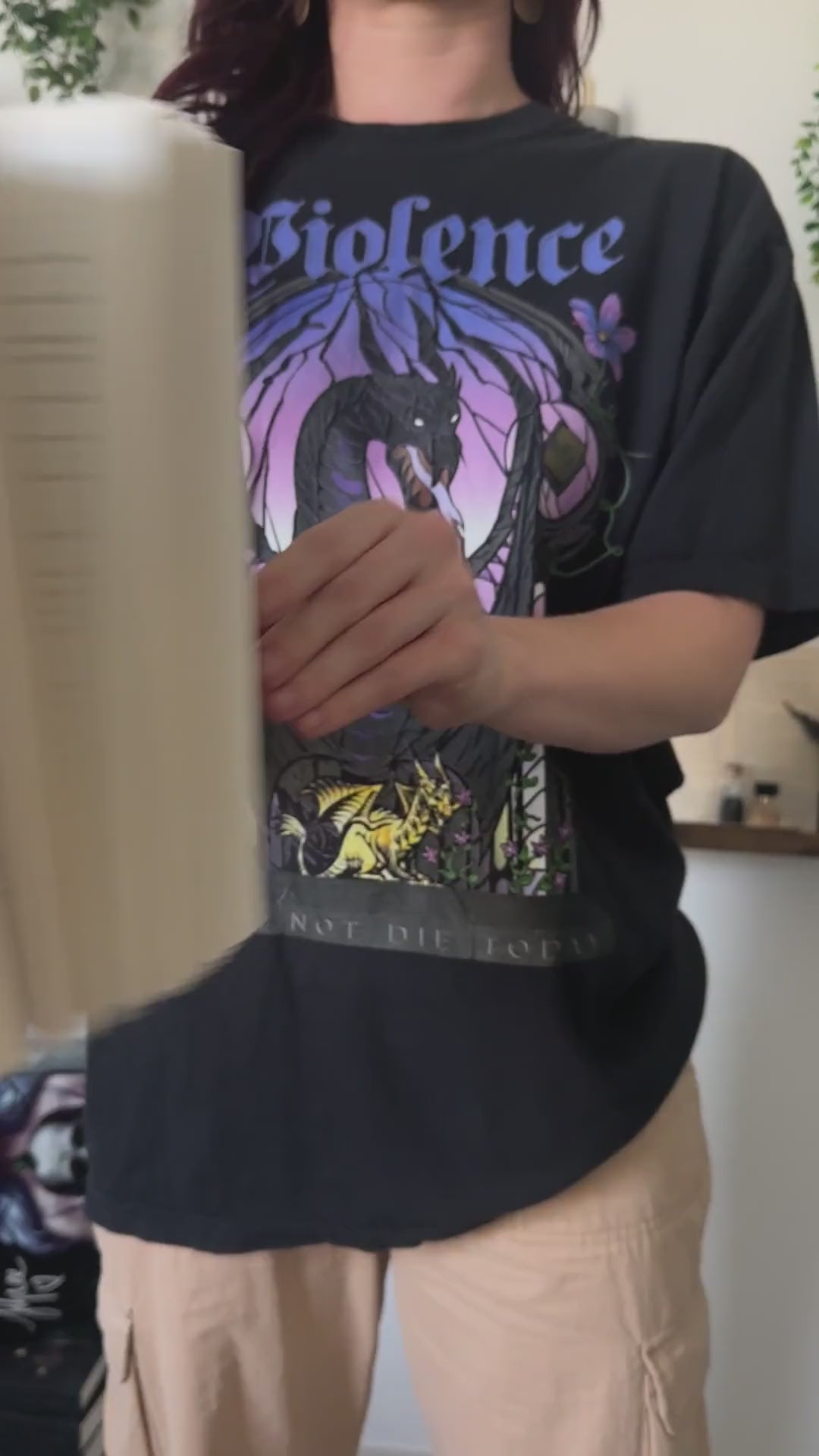Try-on video: the black Violence t-shirt revealed from behind the Fourth Wing paperback — the stained-glass arch in purple and lilac with the dark winged dragon and the golden hatchling at its base [focal 50 35]