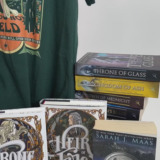 Product video: the forest green Maasverse t-shirt on the hanger with the Throne of Glass hardbacks — the art-nouveau tour poster with 'SJM Presents', MAAS, the World Tour roundel, the three figures in their arches and 'you do not yield' beneath [focal 50 30]