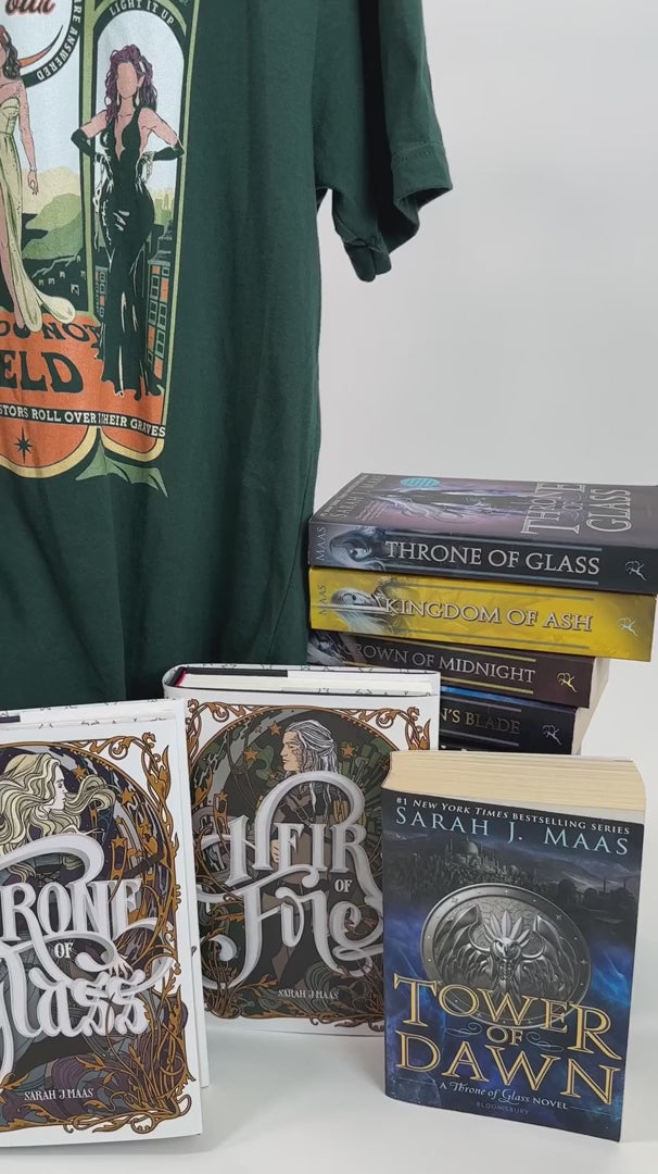 Product video: the forest green Maasverse t-shirt on the hanger with the Throne of Glass hardbacks — the art-nouveau tour poster with 'SJM Presents', MAAS, the World Tour roundel, the three figures in their arches and 'you do not yield' beneath [focal 50 30]