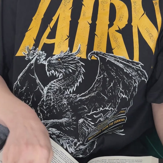 Lifestyle video: the black Tairn t-shirt worn while reading — the gold gothic TAIRN lettering above the white line-art dragon with its wings spread [focal 50 36]