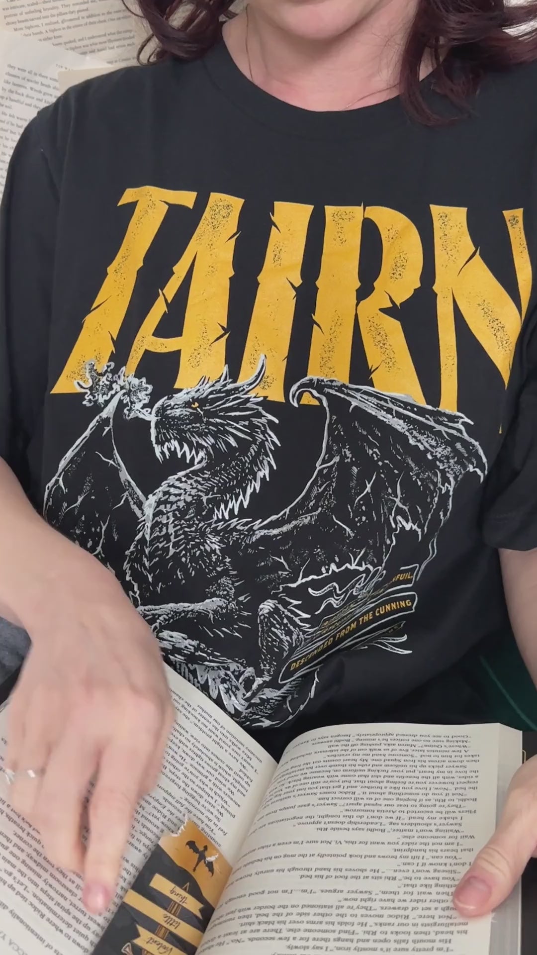Lifestyle video: the black Tairn t-shirt worn while reading — the gold gothic TAIRN lettering above the white line-art dragon with its wings spread [focal 50 36]