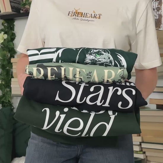Try-on video: the dark green Rowan Whitethorn t-shirt with its vertical WHITETHORN hawk print, shown alongside a folded stack of the other Throne of Glass tees [focal 50 30]