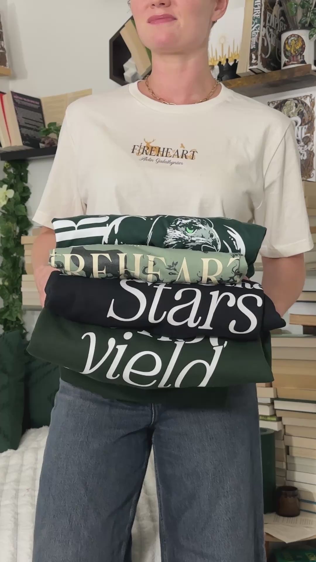 Try-on video: the dark green Rowan Whitethorn t-shirt with its vertical WHITETHORN hawk print, shown alongside a folded stack of the other Throne of Glass tees [focal 50 30]