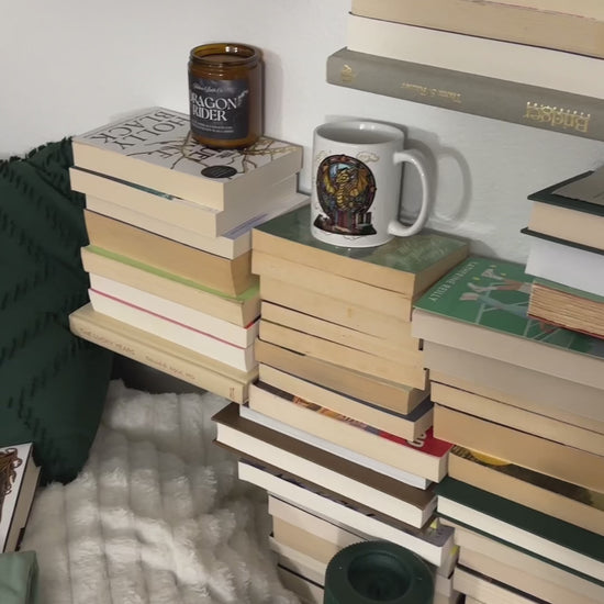 Styled flat lay: the sage and off-white Fireheart tees folded on a bed with Throne of Glass hardbacks, a slow pan across the back print — 'to whatever end', FIREHEART, Aelin Galathynius, 'the world will be saved and remade by the dreamers' [focal 50 60]