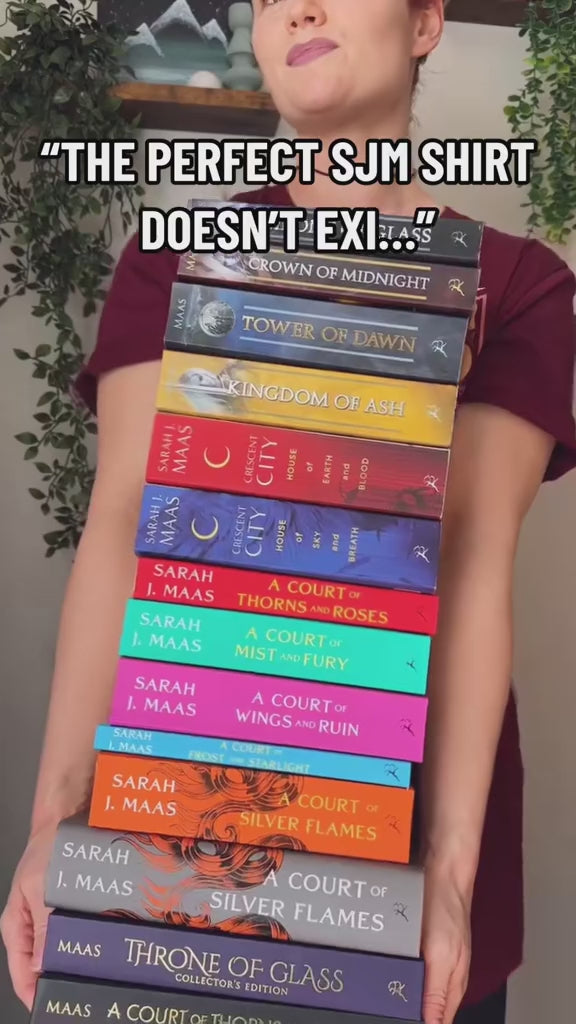 Reveal video: a stack of the whole Sarah J Maas shelf held up over the maroon Maasverse t-shirt, then dropped — the art-nouveau tour poster with MAAS, the World Tour roundel, the three figures in their arches and 'you do not yield' beneath [focal 50 20]