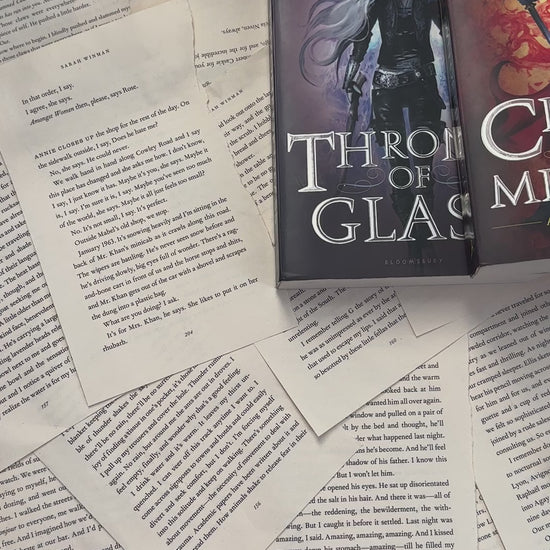 Flat lay video: the black Manon Blackbeak t-shirt laid over scattered book pages with the Throne of Glass, Crown of Midnight and Kingdom of Ash hardbacks — the white tour-poster print reading 'The Thirteen presents / Manon Blackbeak / World Tour 13' around the wyvern [focal 50 50]