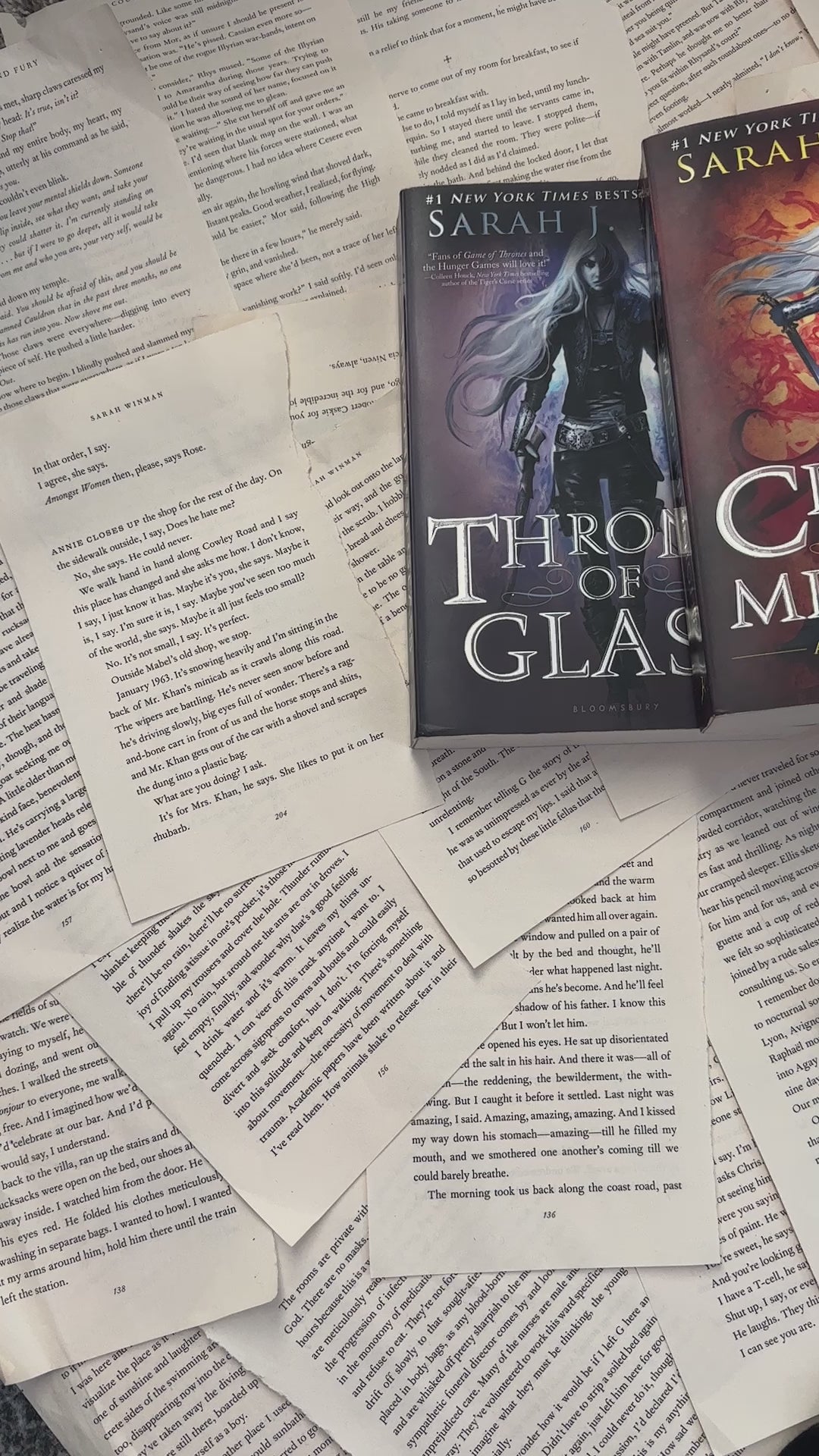 Flat lay video: the black Manon Blackbeak t-shirt laid over scattered book pages with the Throne of Glass, Crown of Midnight and Kingdom of Ash hardbacks — the white tour-poster print reading 'The Thirteen presents / Manon Blackbeak / World Tour 13' around the wyvern [focal 50 50]