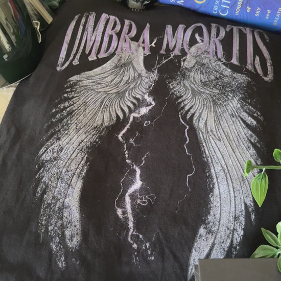 Flat lay video: the black Umbra Mortis t-shirt with the Crescent City books — the arched UMBRA MORTIS lettering above a pair of outstretched wings split by lightning [focal 50 30]