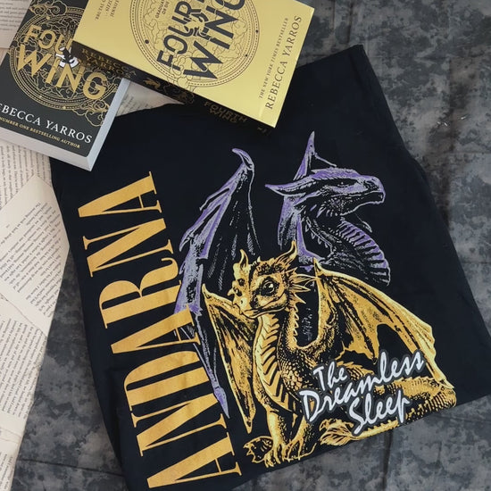 Flat lay video: the black Andarna t-shirt with the Fourth Wing hardback — the gold ANDARNA lettering beside the golden hatchling and the two grey dragons above her [focal 50 55]