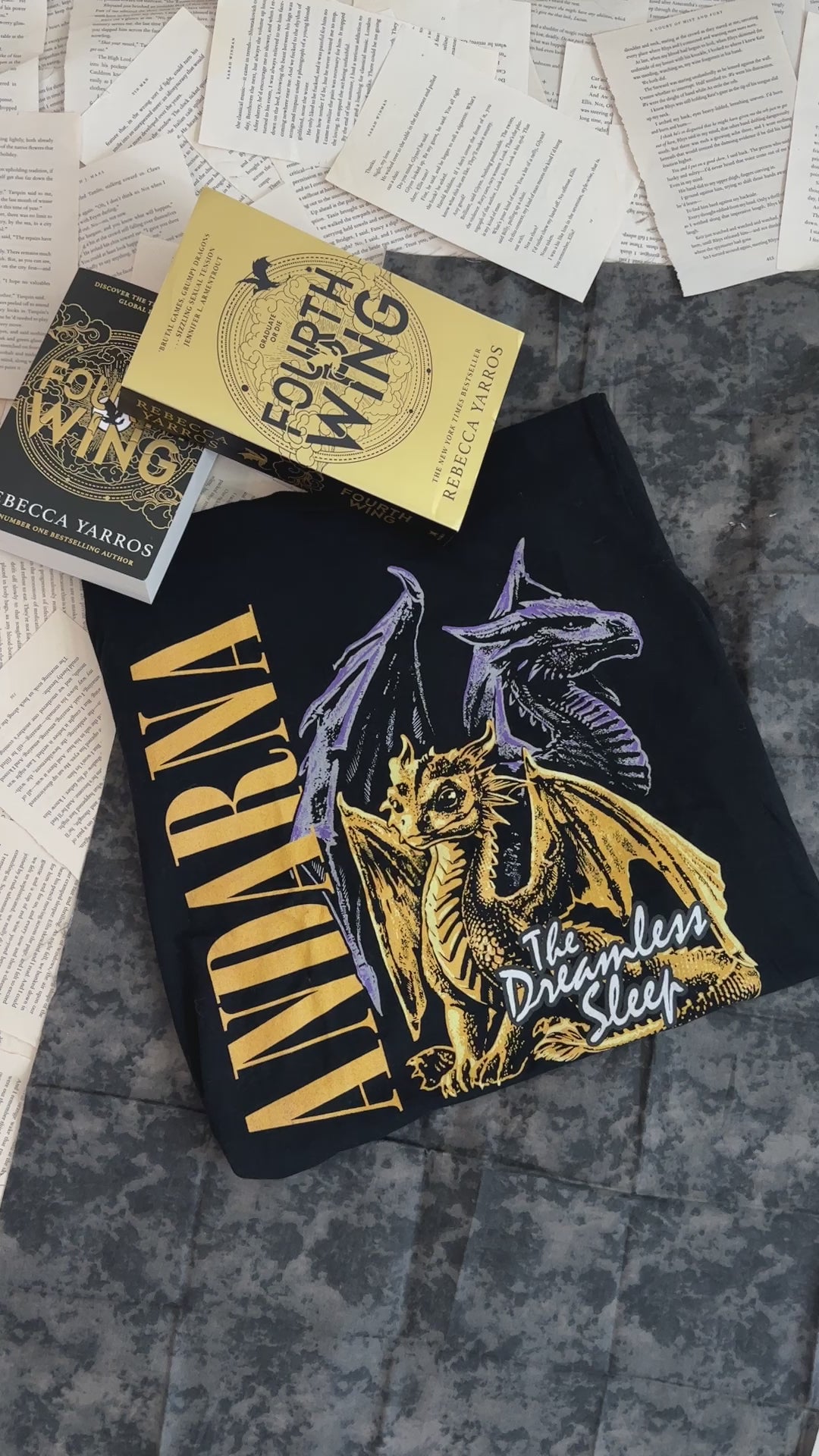 Flat lay video: the black Andarna t-shirt with the Fourth Wing hardback — the gold ANDARNA lettering beside the golden hatchling and the two grey dragons above her [focal 50 55]