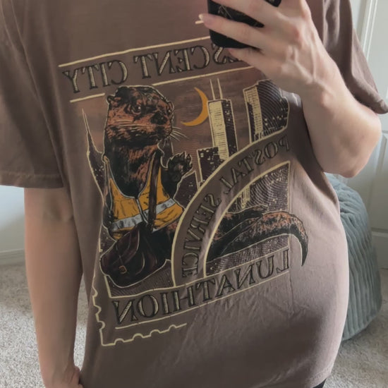Mirror try-on video: oversized taupe Crescent City Otters t-shirt worn with leggings, the arched gold and dark print with the otters filling the chest [focal 50 35]