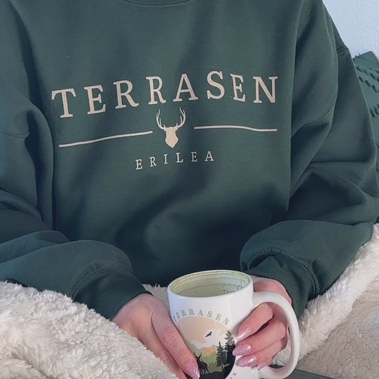 Try-on video: the forest green Terrasen sweatshirt worn under a sherpa blanket with the Terrasen National Park mug — the cream TERRASEN wordmark with the stag head and ERILEA beneath [focal 50 30]