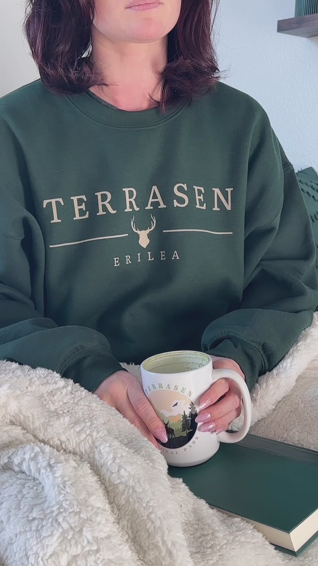 Try-on video: the forest green Terrasen sweatshirt worn under a sherpa blanket with the Terrasen National Park mug — the cream TERRASEN wordmark with the stag head and ERILEA beneath [focal 50 30]