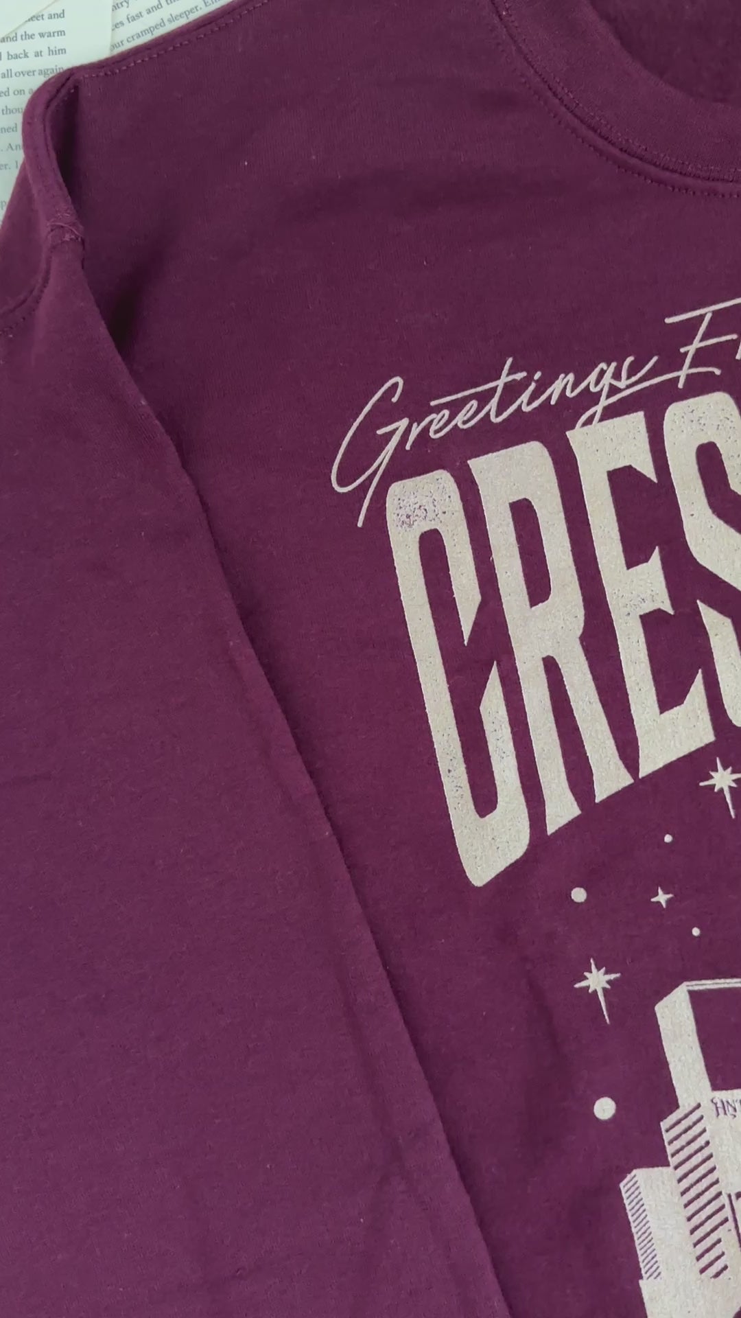 Flat lay video: the maroon Greetings From Crescent City sweatshirt — the blush skyline arch with 'Lunathion' and 'Light it up' beneath the lettering [focal 50 55]