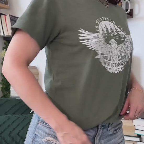 Try-on video: the olive Rowan Whitethorn t-shirt — the circular badge print with 'to whatever end' arched over pines, the eagle with spread wings and WHITETHORN on a banner — then held with a folded stack of the other Throne of Glass tees [focal 50 30]