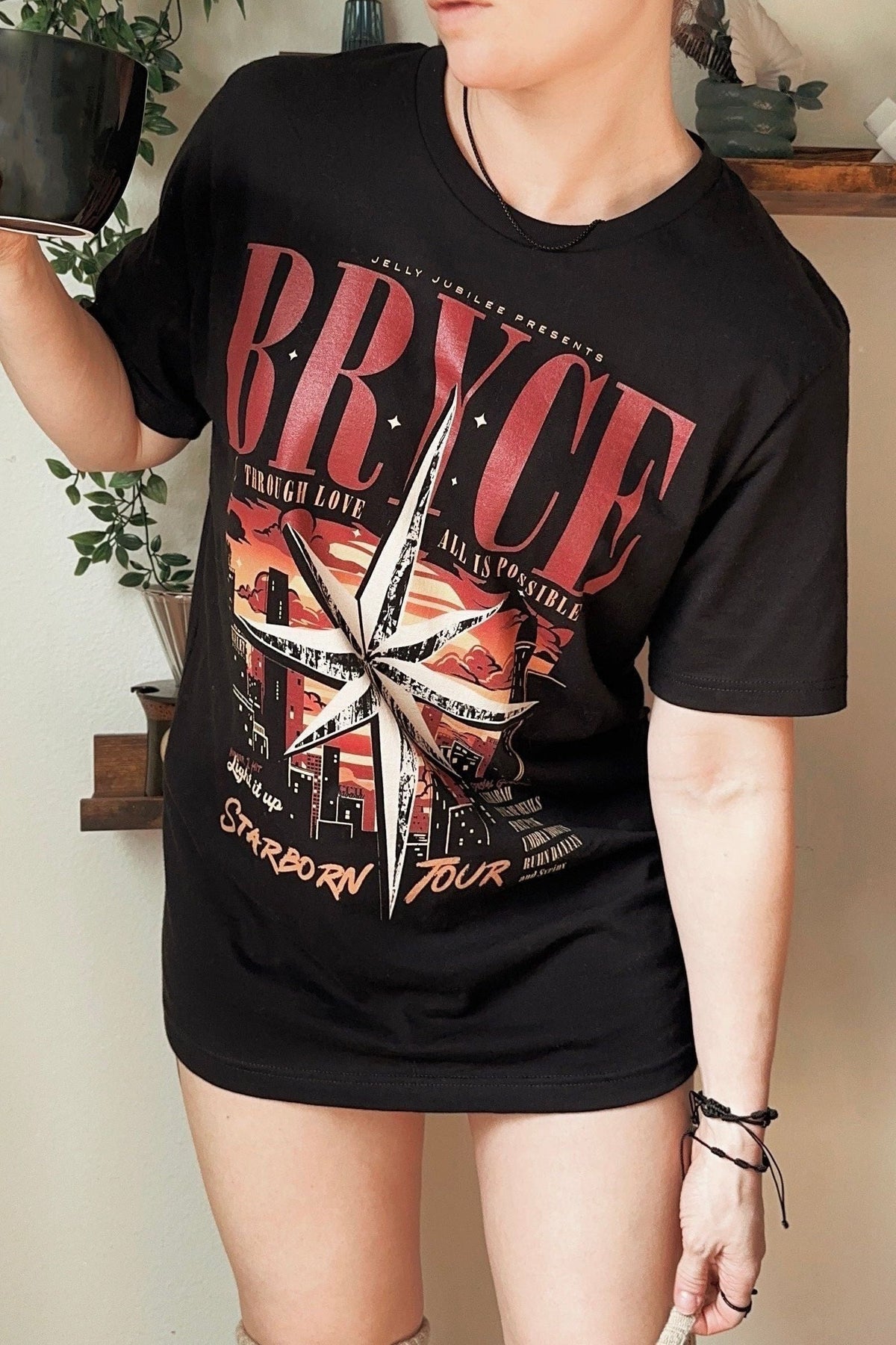 Model wearing a black Bryce t-shirt with a white starburst over a red city skyline and STARBORN TOUR lettering, inspired by Crescent City by Sarah J. Maas