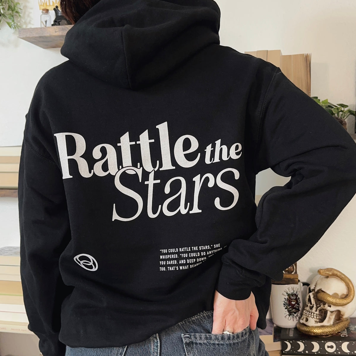 Back view of a black hoodie printed with Rattle the Stars in white serif type above a small quote block, inspired by Throne of Glass by Sarah J. Maas