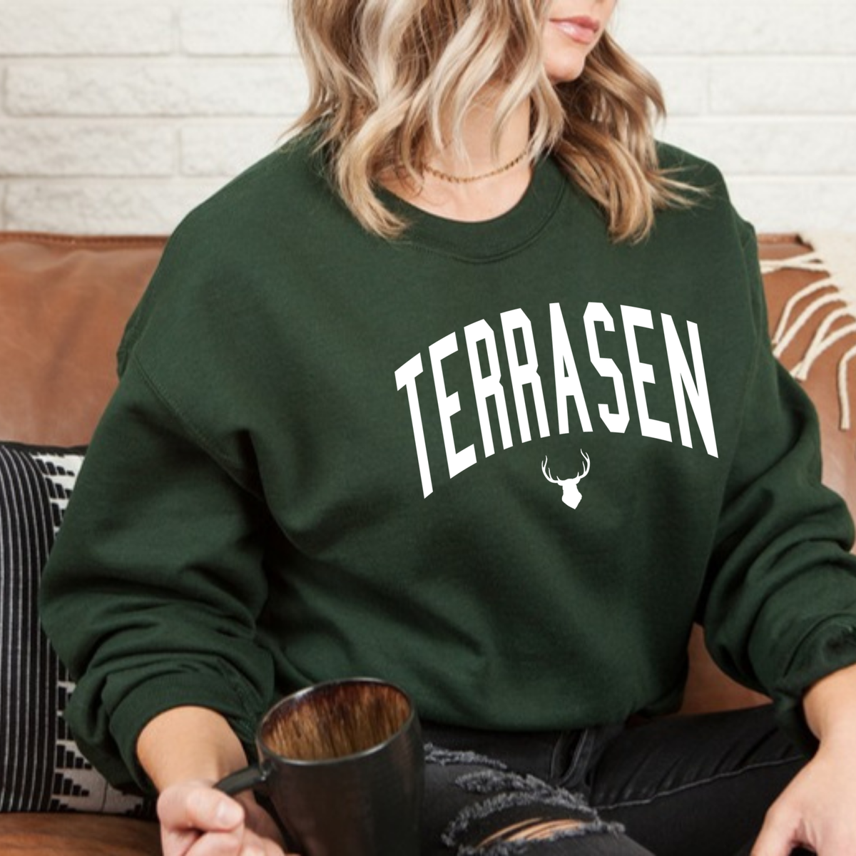 Model on a leather sofa in a forest green Terrasen sweatshirt with white arched lettering and a stag, inspired by Throne of Glass by Sarah J. Maas