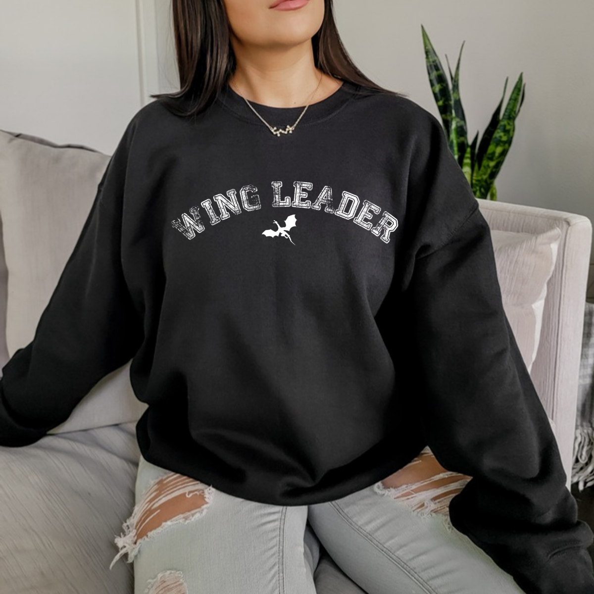Model seated on a sofa in a black Wing Leader sweatshirt, inspired by Throne of Glass by Sarah J. Maas